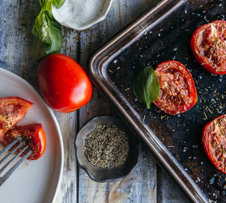 Au der Vogelperspektive: Auf einem Ofenblech liegen halbierte Tomaten, die mit Kräutern betreut sind. Daneben steht ein weißer Teller, auf dem ebenfalls eine halbierte Tomate liegt. | © Shutterstock / Karen Hibbard Photography