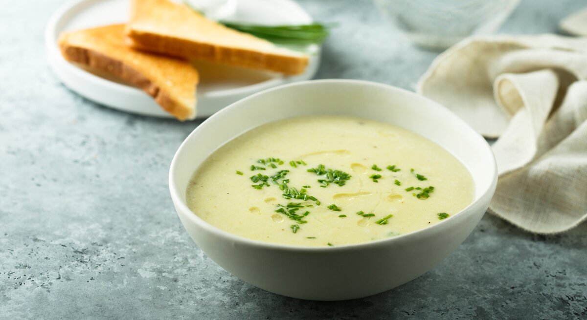 Eine weiße Schale mit der grünen Suppe auf bläulichem Untergrund. Alles von der Seite fotografiert.