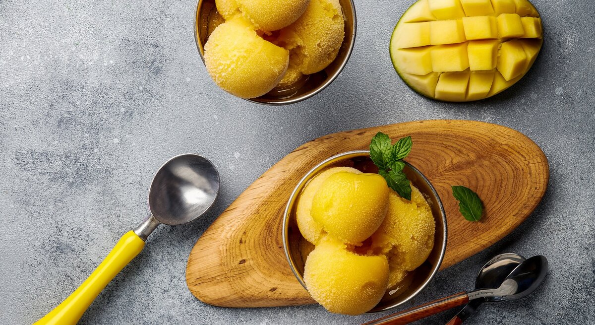 Ein paar Kugeln Mango-Joghurt-Eis in zwei Schälchen auf einem grauen Tisch. Daneben eine eingeritzte Mangohälfte und ein paar Löffel. Alles direkt von oben fotografiert.