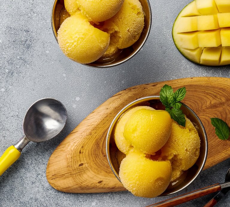 Ein paar Kugeln Mango-Joghurt-Eis in zwei Schälchen auf einem grauen Tisch. Daneben eine eingeritzte Mangohälfte und ein paar Löffel. Alles direkt von oben fotografiert. | © Shutterstock/ Tanya Sid