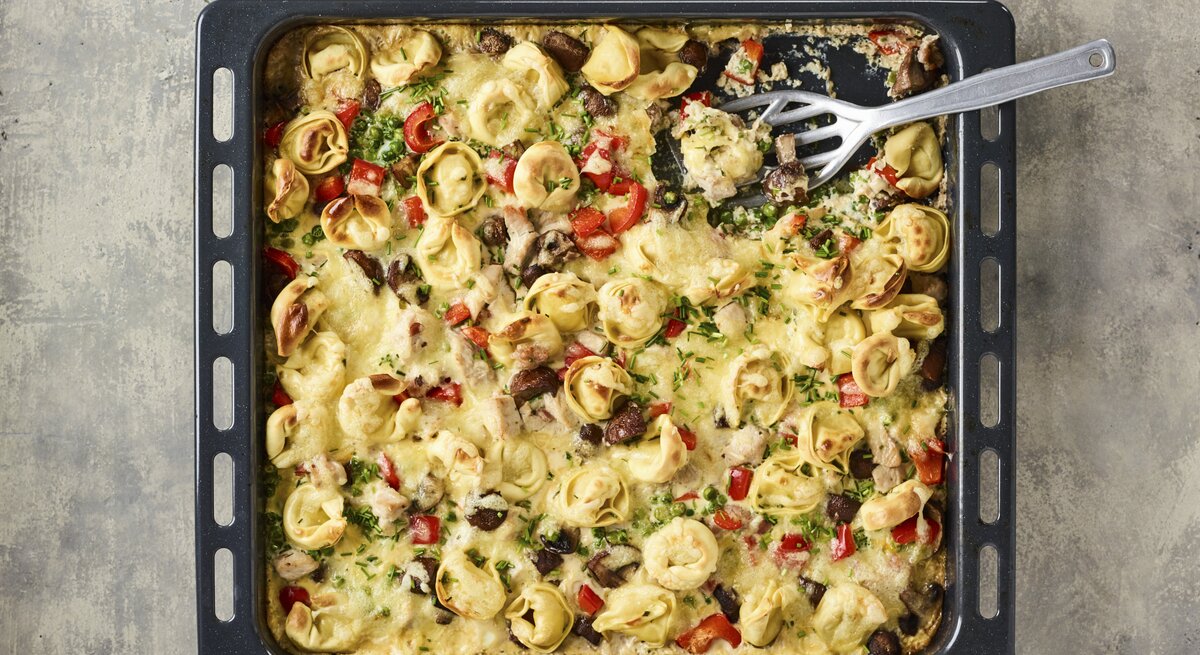 Überbackener Tortelloni-Auflauf auf Backblech mit knackiger Paprika, Kräutern, knusprigem Speck in cremiger Sahnesoße auf steinernem Untergrund