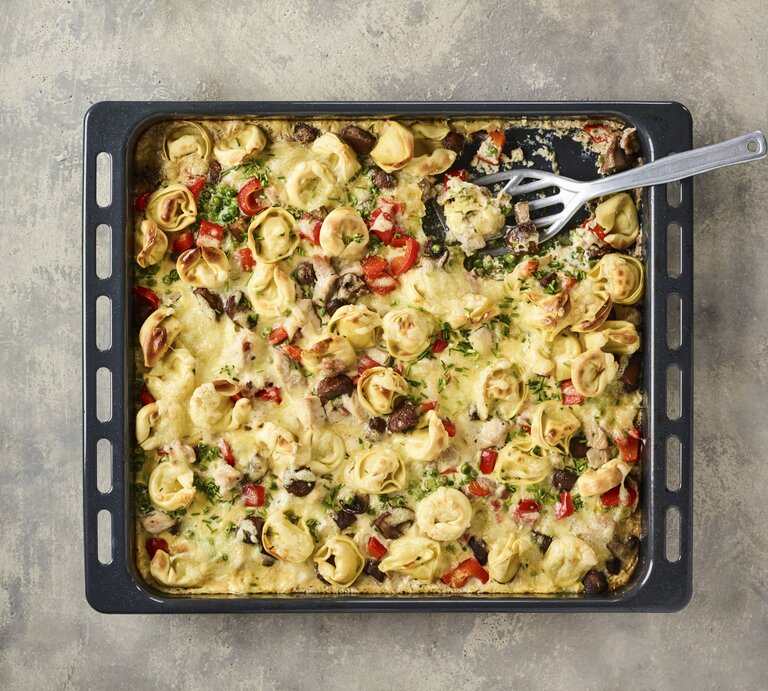 Überbackener Tortelloni-Auflauf auf Backblech mit knackiger Paprika, Kräutern, knusprigem Speck in cremiger Sahnesoße auf steinernem Untergrund | © Stockfood/Gräfe & Unzer Verlag / Arras, Klaus