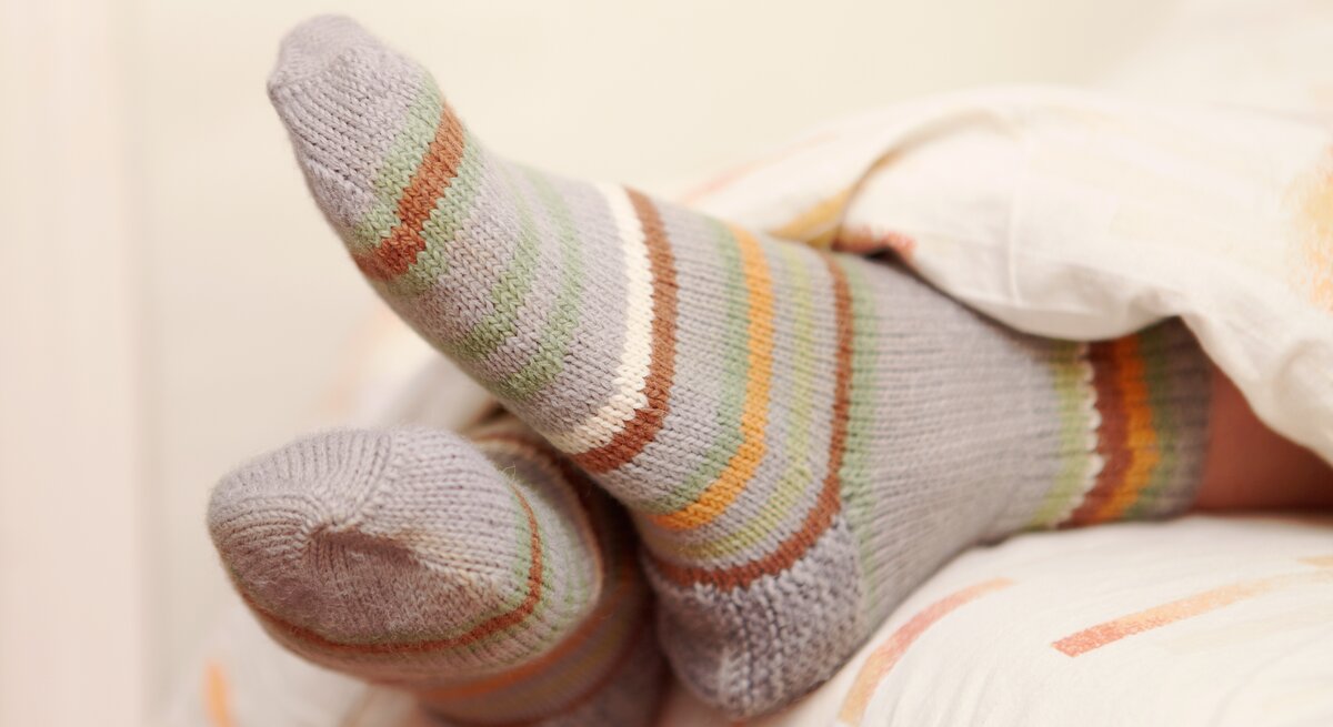 Eine Person liegt entspannt im Bett mit den Füßen hoch, die in gestreiften Socken in sanften Grau-, Grün-, Orange- und Weißtönen gehüllt sind. Der Fokus liegt auf den gemütlichen Socken, die gegen die weiche Bettwäsche drücken.
