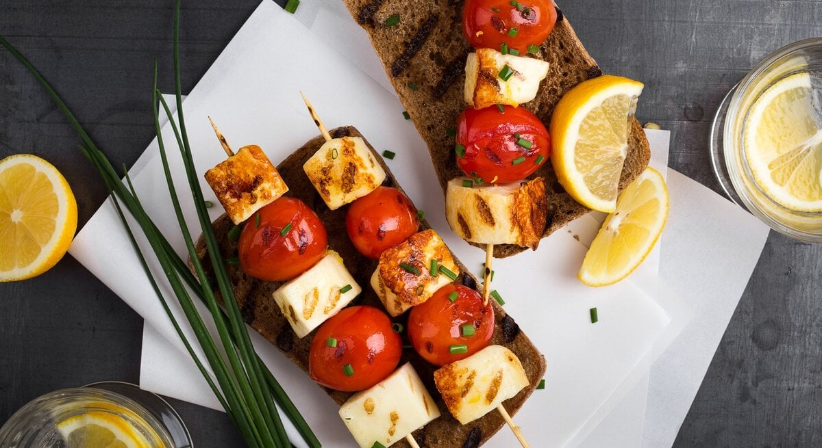 Halloumi-Tomaten-Spieße auf Brotscheiben. Daneben Zitrone und Schnittlauch. 