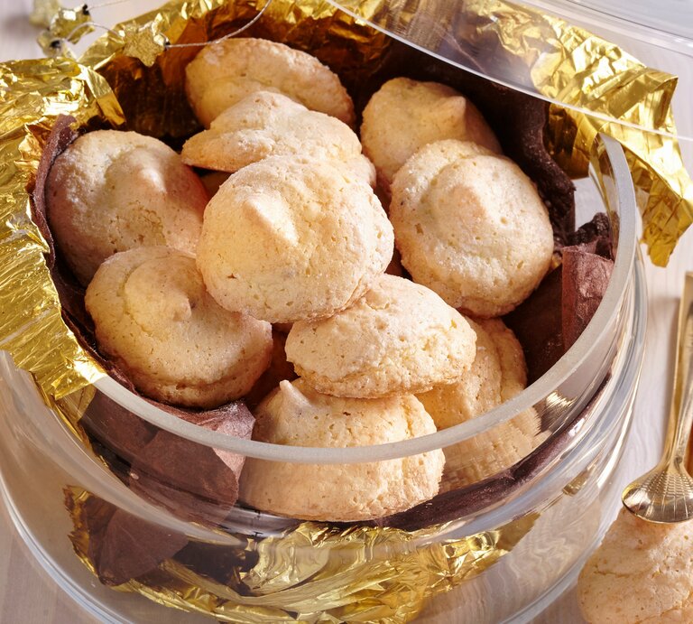 Mehrere Zitronen-Makronen liegen in einer Schale aus Glas. Die Makronen sind umgeben von goldener Folie. Einige Schokoladenstückchen liegen zwischen den Makronen. | © Stockfood / Profimedia