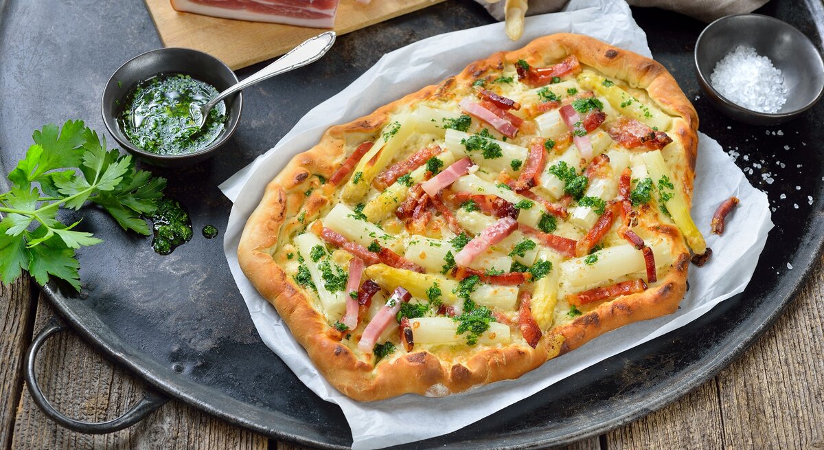 Spargel-Flammkuchen auf einem Stück Backpapier. Daneben verschiedene Zutaten.