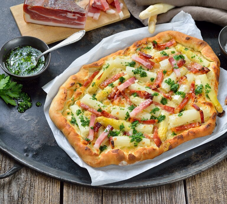 Spargel-Flammkuchen auf einem Stück Backpapier. Daneben verschiedene Zutaten. | © Getty Images / kabVisio