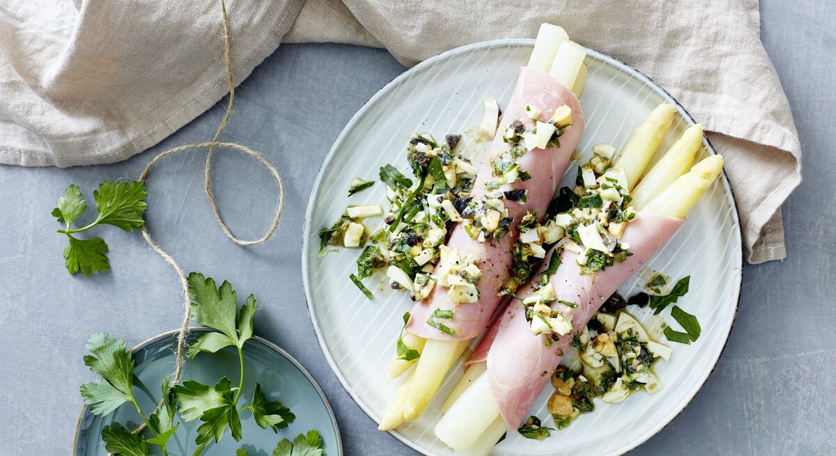 Mediterraner Spargel eingewickelt in Schinken auf einem hellblauen Teller neben Petersilie auf einem gedeckten hellblauen Tisch