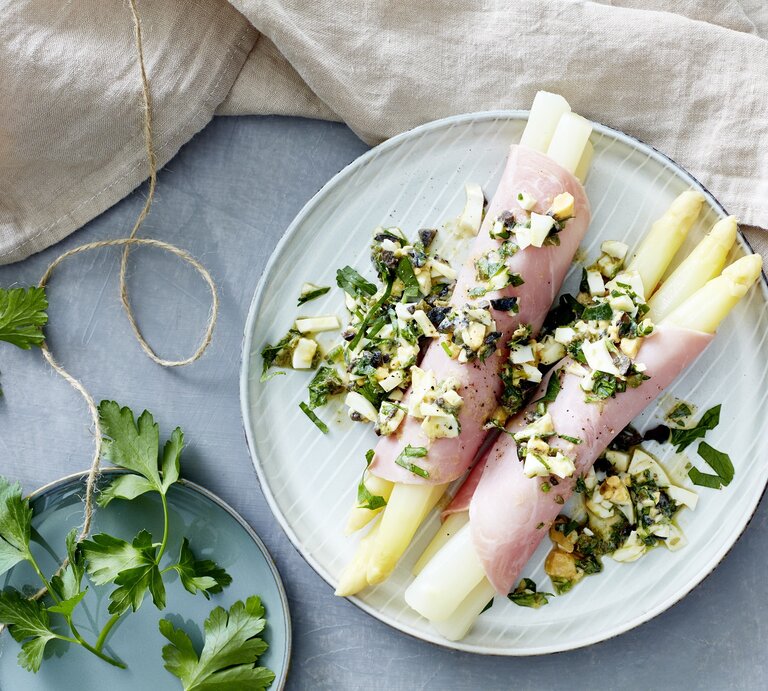 Mediterraner Spargel eingewickelt in Schinken auf einem hellblauen Teller neben Petersilie auf einem gedeckten hellblauen Tisch | © EAT CLUB / Isabell Triemer