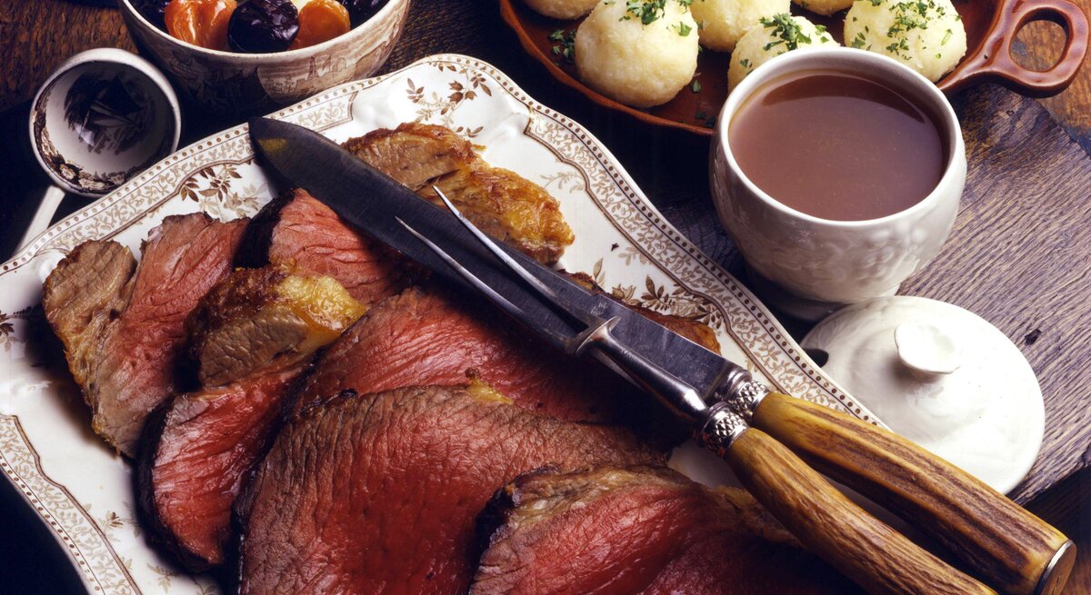 Angeschnittener Sauerbraten auf einem weißen Servierteller, daneben steht eine schale voll Klöße.