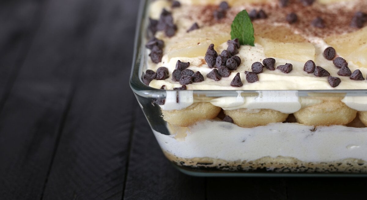 Gläserne Auflaufform gefüllt mit Ananas-Tiramisu auf dunklem Untergrund aus Holz. 