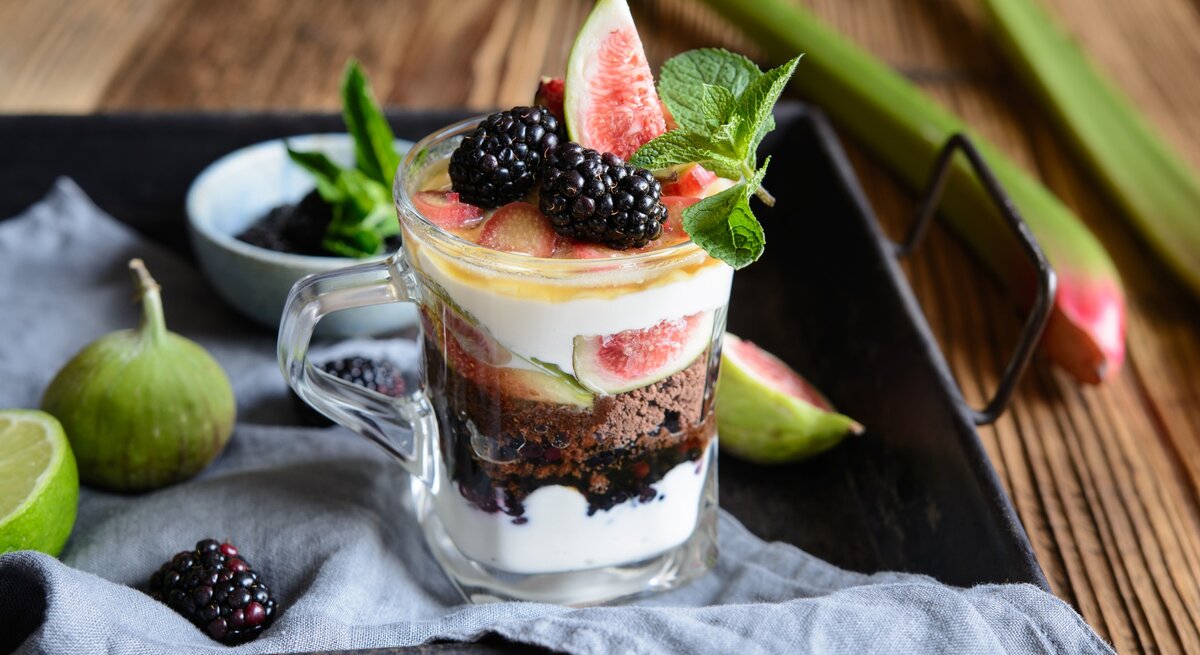 Schichtdessert mit Rhabarber, Feigen, Ricottacreme, Kaffeelikörkeksen Brombeeren und Minzblatt auf schwarzem Tablette mit Beeren und Feigen. Daneben auf Holztische Rhabarber. Frontalansicht.