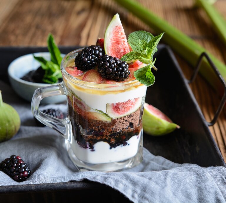 Schichtdessert mit Rhabarber, Feigen, Ricottacreme, Kaffeelikörkeksen Brombeeren und Minzblatt auf schwarzem Tablette mit Beeren und Feigen. Daneben auf Holztische Rhabarber. Frontalansicht. | © Getty Images / NoirChocolate