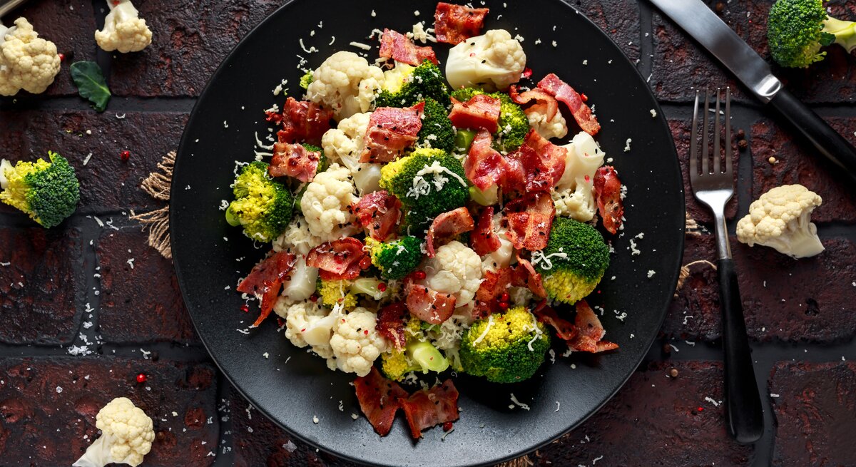 Ein Teller Blumenkohl-Brokkoli-Salat mit Parmaschinken, Parmesan und Sesam, drumherum liegen Kohlröschen und Besteck.