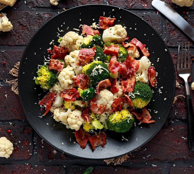 Ein Teller Blumenkohl-Brokkoli-Salat mit Parmaschinken, Parmesan und Sesam, drumherum liegen Kohlröschen und Besteck. | © Getty Images/ DronG