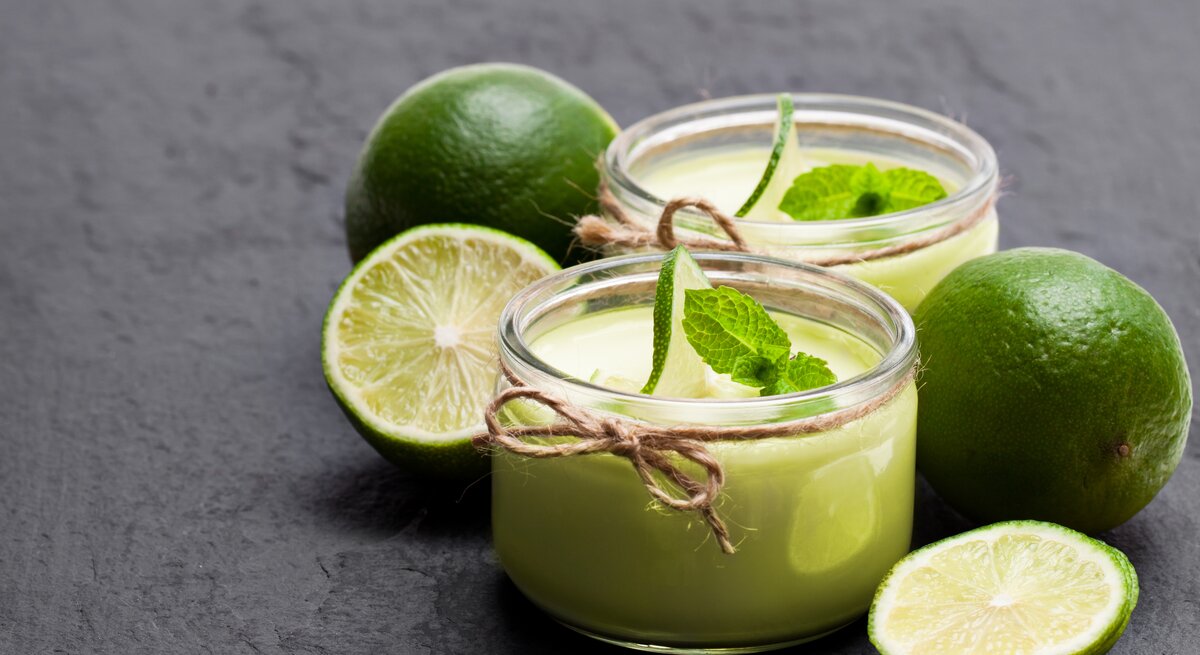 Frontalsicht: Auf einem Schiefer-Untergrund stehen zwei Gläser Limetten-Joghurt mit Minze. Drum herum liegen zwei ganze Limetten und zwei Limettenhälften.