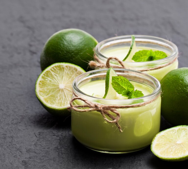 Frontalsicht: Auf einem Schiefer-Untergrund stehen zwei Gläser Limetten-Joghurt mit Minze. Drum herum liegen zwei ganze Limetten und zwei Limettenhälften. | © Shutterstock / Elena Zajchikova
