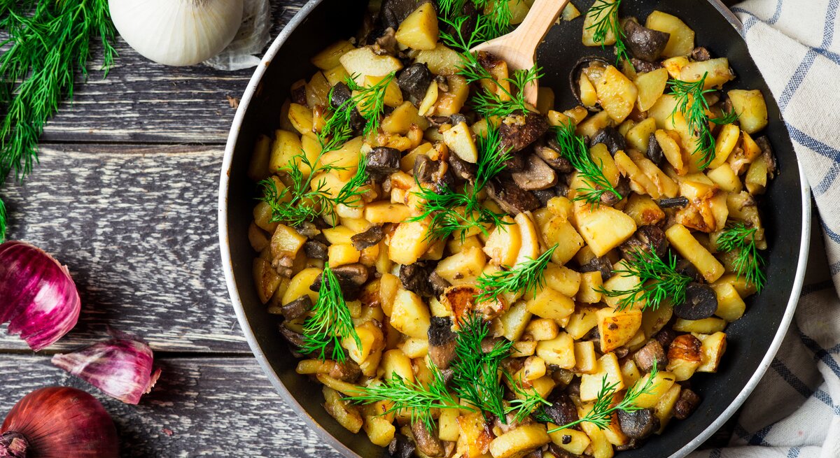 Draufsicht: Eine Pfanne mit Bratkartoffel mit Birne und Pilzen und Dill steht auf Holz. Naneben liegen eine weiße und eine rote Zwiebel sowie frischer Dill.