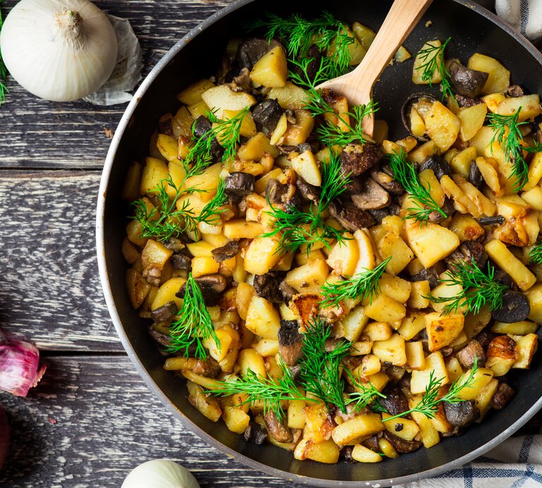 Draufsicht: Eine Pfanne mit Bratkartoffel mit Birne und Pilzen und Dill steht auf Holz. Naneben liegen eine weiße und eine rote Zwiebel sowie frischer Dill. | © Shutterstock /  Dragon_Fly