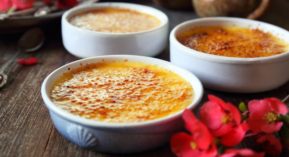 Drei Eierlikör-Crème brûlée Schalen auf einem dunklen holztisch mit roten Blüten, seitlich fotografiert.