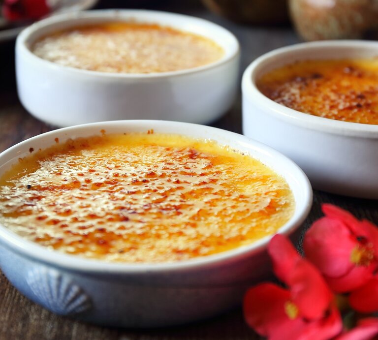 Drei Eierlikör-Crème brûlée Schalen auf einem dunklen holztisch mit roten Blüten, seitlich fotografiert. | © Shutterstock/photosimysia