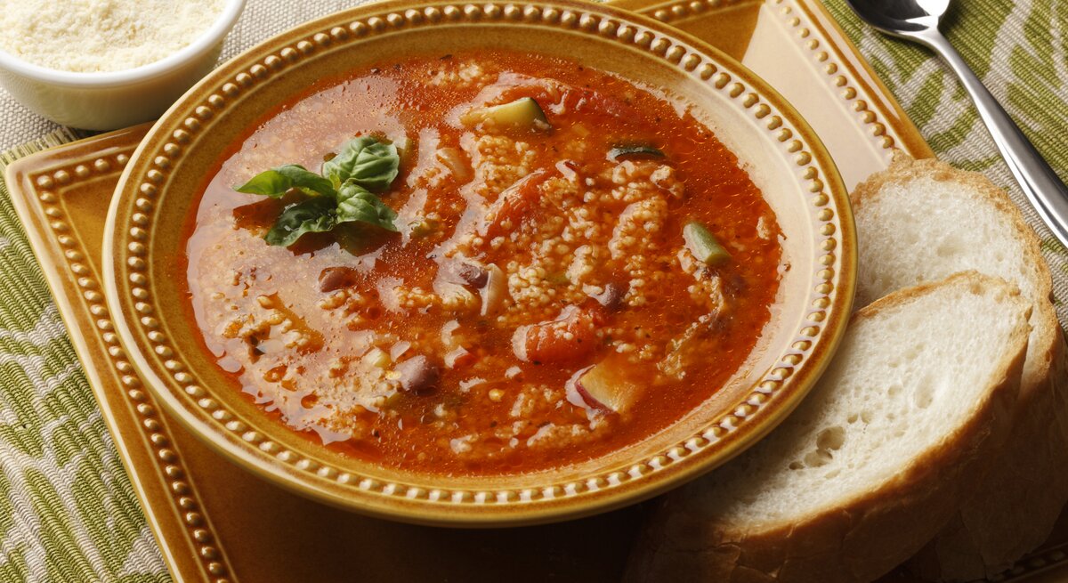 Ein Teller Couscous-Suppe mit Zucchini und Tomate steht auf einem Tablett. Daneben lieben zwei Scheiben Brot.