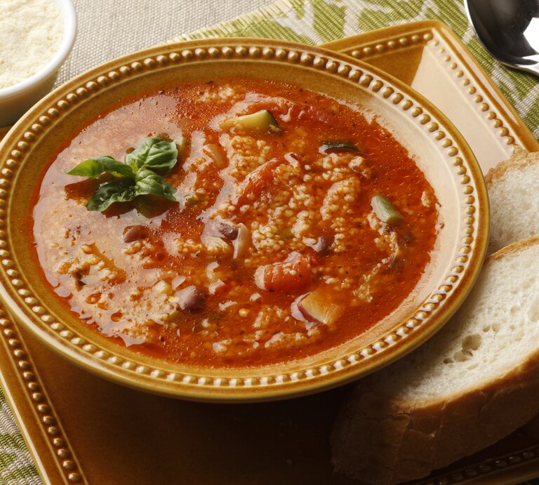 Ein Teller Couscous-Suppe mit Zucchini und Tomate steht auf einem Tablett. Daneben lieben zwei Scheiben Brot. | © Getty Images; Tim Hawley