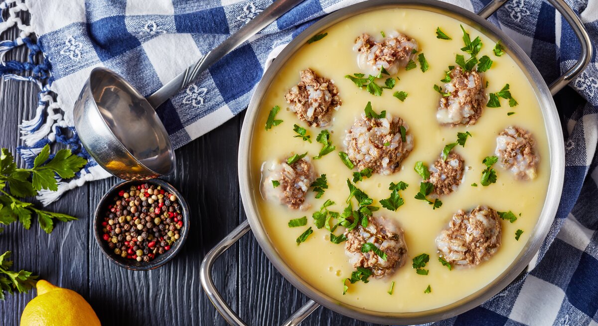 Griechische Hackbällchensuppe mit Zitrone in einem Topg aus Edelstahl neben einer Suppenkelle, Pfefferkörnern und frischer Zitrone auf einem blauen Holztisch mit blau karierter Tischdecke.