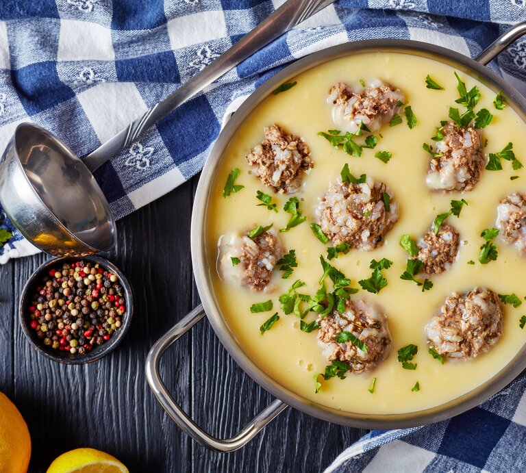 Griechische Hackbällchensuppe mit Zitrone in einem Topg aus Edelstahl neben einer Suppenkelle, Pfefferkörnern und frischer Zitrone auf einem blauen Holztisch mit blau karierter Tischdecke. | © Getty Images / from_my_point_of_view