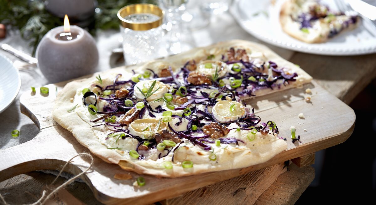 Ein Rotkohl-Flammkuchen mit Ziegenkäse liegt auf einem Holzbrett auf einem Holztisch. Im Hintergrund stehen Gläser und mehrere Kerzen. Der Flammkuchen ist angerichtet mit Walnüssen und Frühlingszwiebelringen.