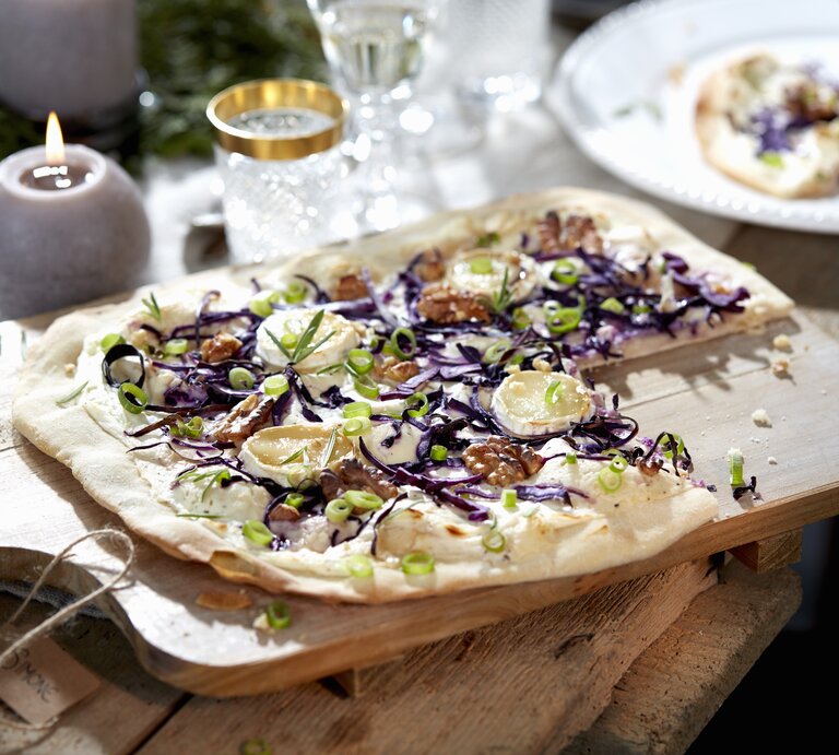 Ein Rotkohl-Flammkuchen mit Ziegenkäse liegt auf einem Holzbrett auf einem Holztisch. Im Hintergrund stehen Gläser und mehrere Kerzen. Der Flammkuchen ist angerichtet mit Walnüssen und Frühlingszwiebelringen. | © Stockfood / Przibylla, Dirk