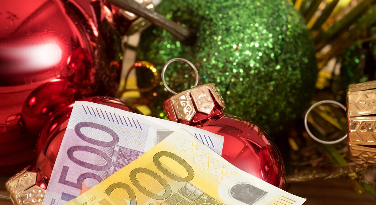 Ein 200-Euro- und ein 500-Euro-Schein liegen vor Weihnachtsdeko. Im Hintergrund sind ein grüner Deko-Apfel und Christbaumkugeln zu sehen.