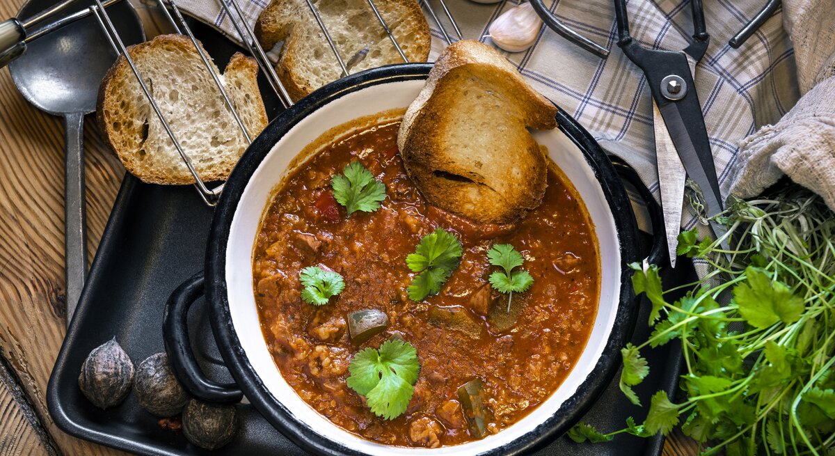 Auf einem Holztisch liegen Brotschreiben, eine Schere, frische Kräuter und ein Suppenlöffel. Darüber hinaus steht dort ein Topf Lammsuppe aus Georgien. Das Gericht nennt man Kharcho.