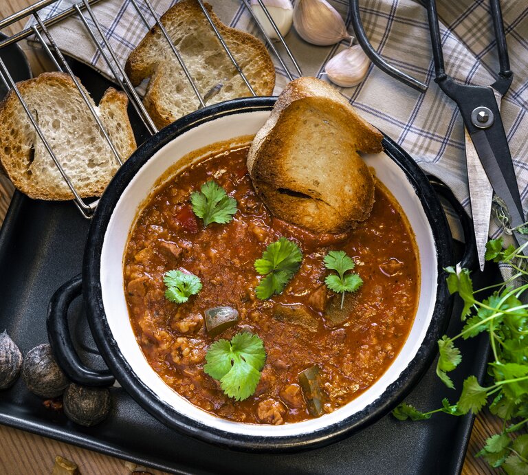 Auf einem Holztisch liegen Brotschreiben, eine Schere, frische Kräuter und ein Suppenlöffel. Darüber hinaus steht dort ein Topf Lammsuppe aus Georgien. Das Gericht nennt man Kharcho. | © Getty Images / Jacek Kadaj