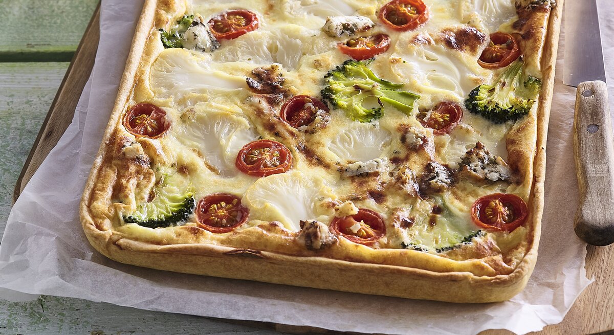 Zu sehen ist ein Blechkuchen mit Tomaten, Blumenkohl, Brokkoli, Wurst und überbackenem Käse in der Draufsicht auf Backpapier und Holzbrett. Rechts Messer mit Holzgriff. Alles liet auf grün-braunem Tisch mit einzelnen Brettern.  