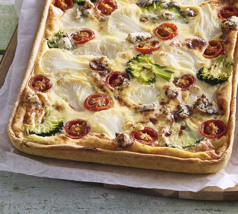 Zu sehen ist ein Blechkuchen mit Tomaten, Blumenkohl, Brokkoli, Wurst und überbackenem Käse in der Draufsicht auf Backpapier und Holzbrett. Rechts Messer mit Holzgriff. Alles liet auf grün-braunem Tisch mit einzelnen Brettern.   | © StockFood / Profimedia