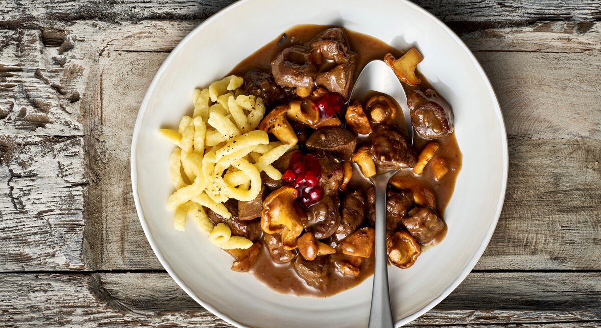 Ein weißer Teller mit einer Portion des Rehgulaschs mit Spätzle und Preiselbeeren, verfeinert mit Pfifferlingen. Ein Löffel ist auf dem Teller platziert, der vor einem Holzhintergrund von oben dargestellt ist.  
