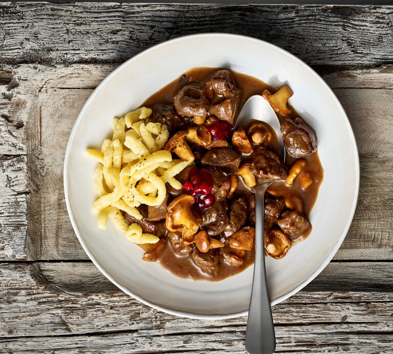 Ein weißer Teller mit einer Portion des Rehgulaschs mit Spätzle und Preiselbeeren, verfeinert mit Pfifferlingen. Ein Löffel ist auf dem Teller platziert, der vor einem Holzhintergrund von oben dargestellt ist.   | © StockFood / Gräfe & Unzer Verlag / Einwanger, Klaus-Maria