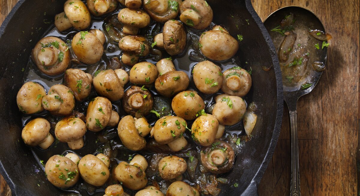 Das Bild zeigt eine gusseiserne Pfanne mit gebratenen Champignons, die auf einem rustikalen Holztisch steht. Die Pilze sehen saftig aus und sind mit Kräutern, wahrscheinlich Petersilie, bestreut
