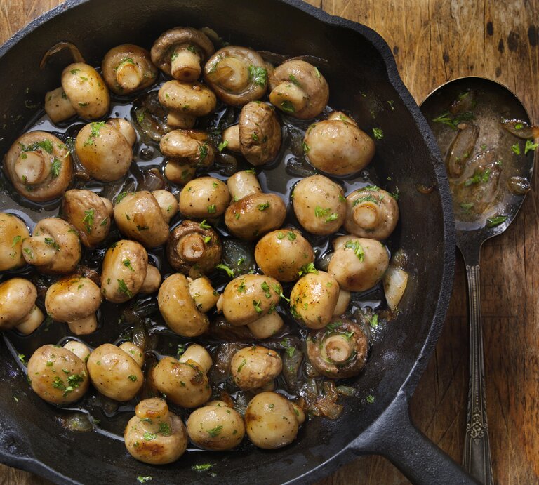 Das Bild zeigt eine gusseiserne Pfanne mit gebratenen Champignons, die auf einem rustikalen Holztisch steht. Die Pilze sehen saftig aus und sind mit Kräutern, wahrscheinlich Petersilie, bestreut | © iStock.com/LauriPatterson