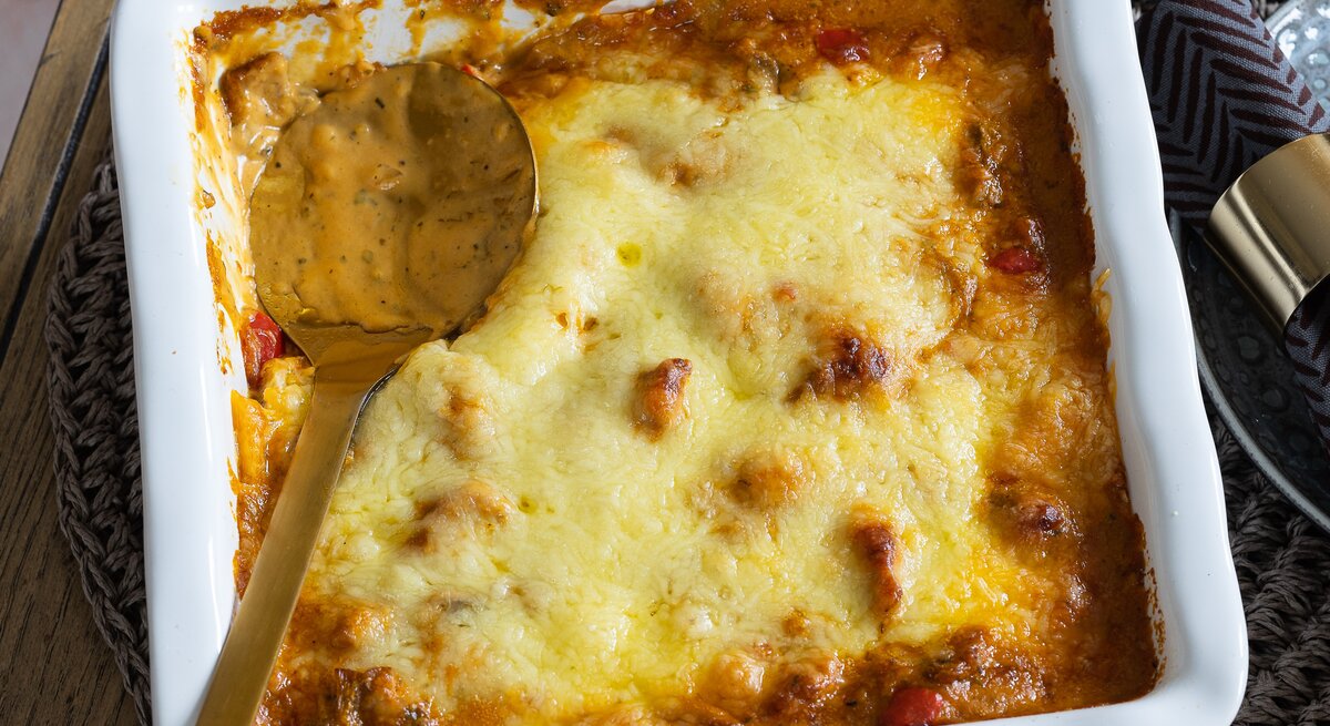 Eine Auflaufform mit Gyros-Auflauf mit Metaxasoße, darin ein großer Löffel zum Auffüllen.
