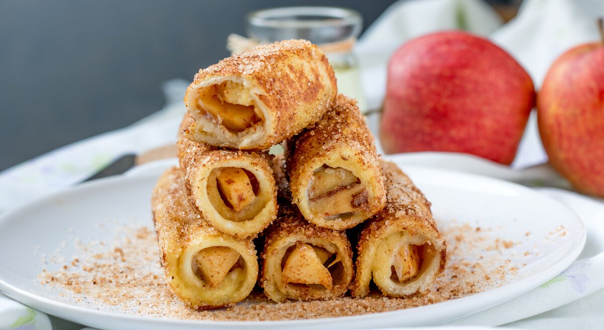 Sechs übereinandergestapelte Apfel-Toast-Röllchen mit Zimt und Zucker bestreut auf einem weißen Teller mit Äpfeln im Hintergrund.