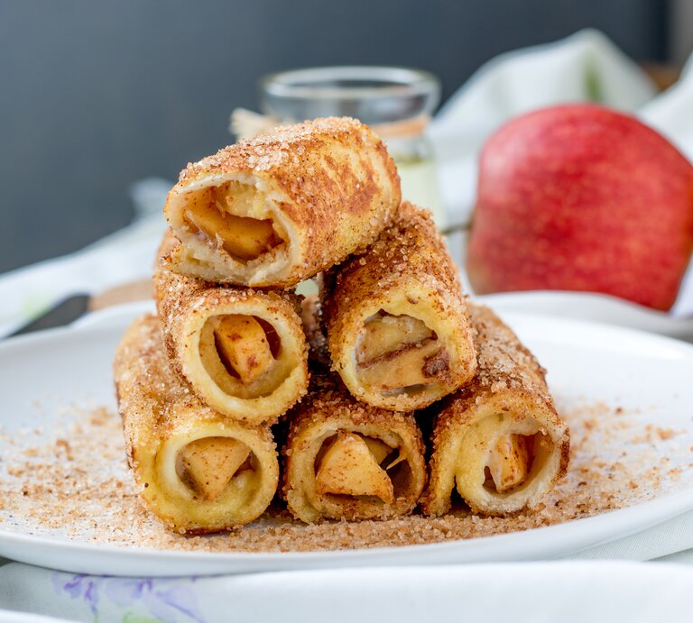 Sechs übereinandergestapelte Apfel-Toast-Röllchen mit Zimt und Zucker bestreut auf einem weißen Teller mit Äpfeln im Hintergrund. | © Shutterstock / Aris Setya