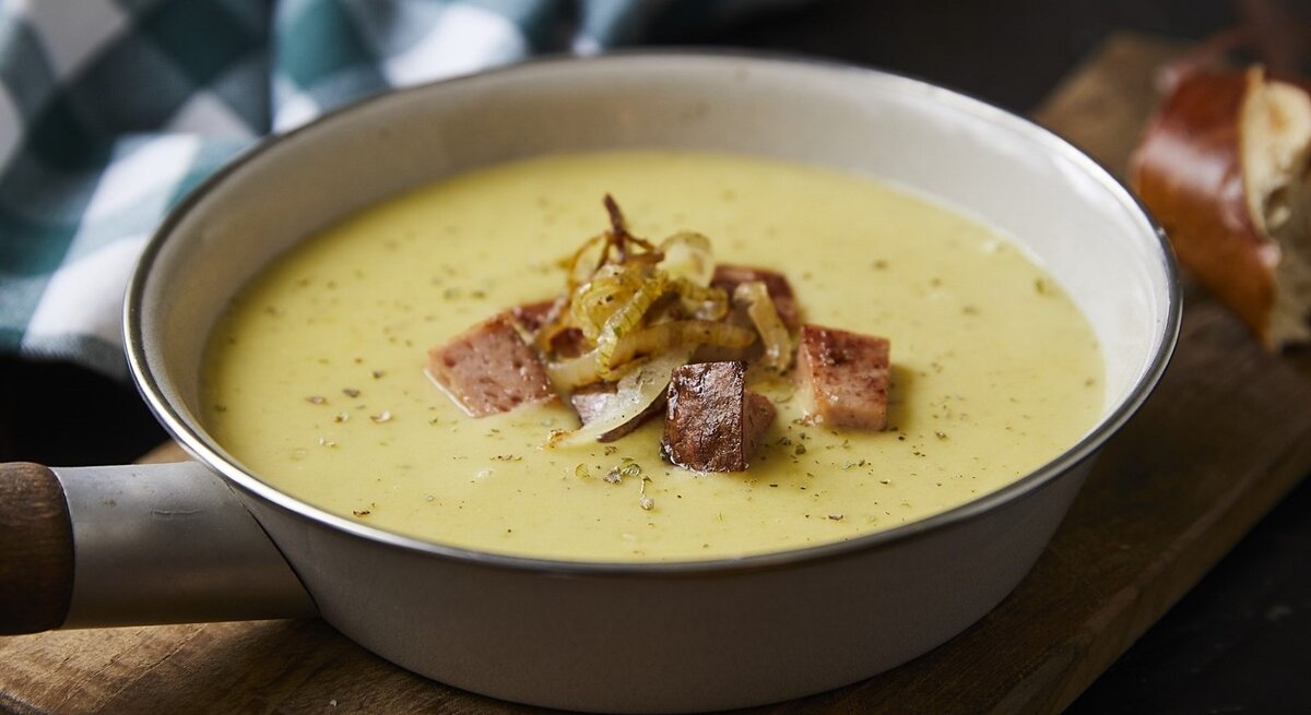 Auf einem Tisch steht eine Pfanne mit einer Kartoffelsuppe mit gebratenem Leberkäse. Sie sieht würzig und aromatisch aus. 