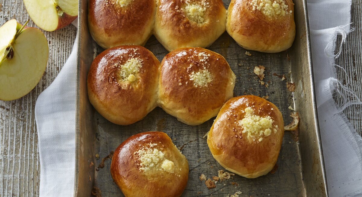 Auf einem Backblech liegen einige Hefebrötchen mit Apfel. Sie sehen fluffig und weich aus. Daneben sind aufgeschnittene Äpfel zu sehen. 