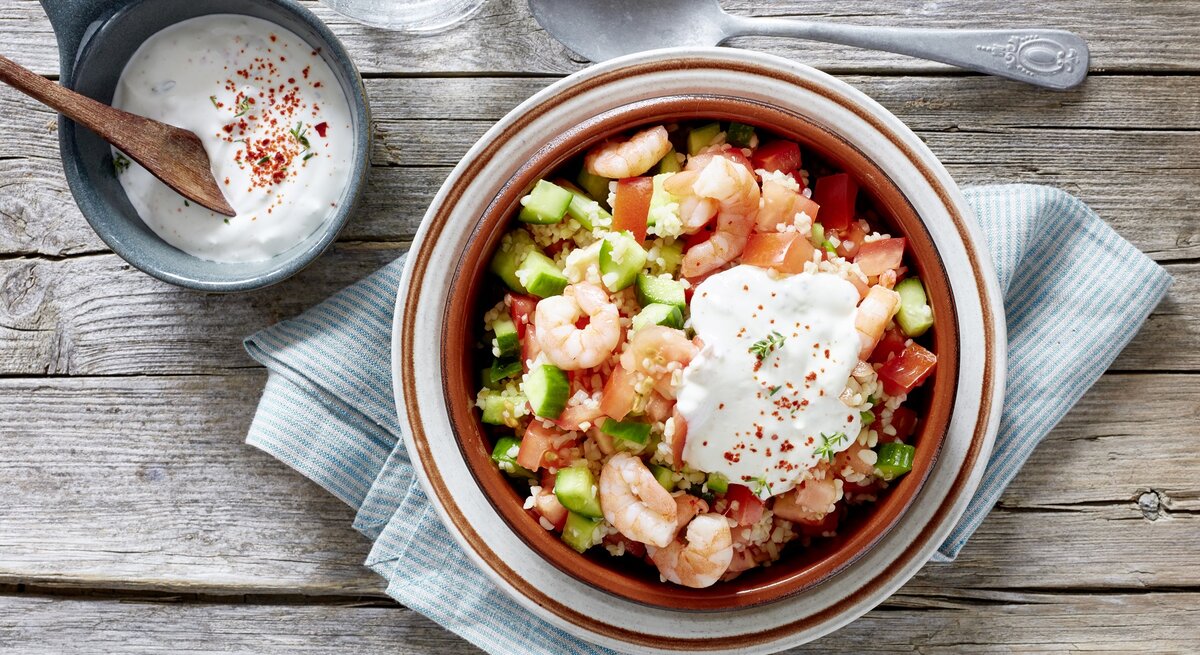 Bulgur-Salat mit Garnelen, Tomaten, Gurken und frischem Joghurt-Dip, serviert auf einem rustikalen Holztisch, von oben fotografiert. 