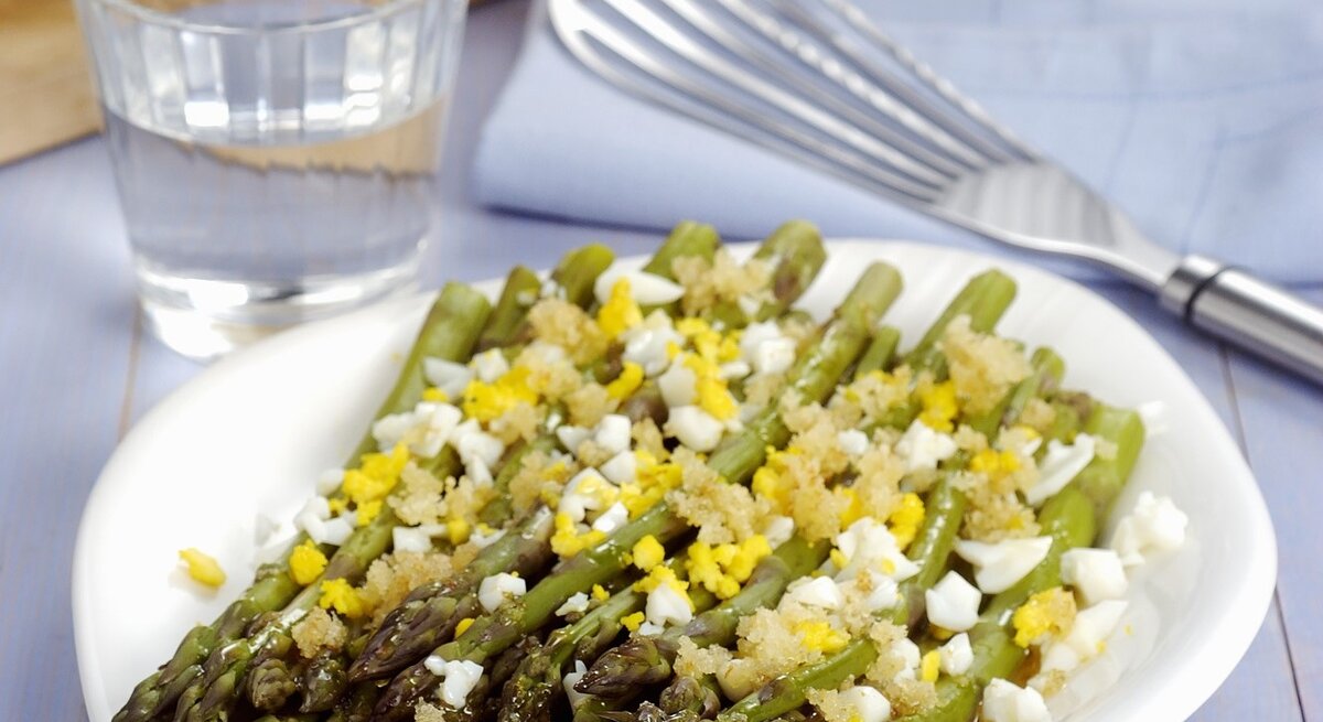 Grüner Spargel auf polnische Art, serviert mit weich gekochtem Ei und einer feinen Estragon-Vinaigrette. Alles auf weißem Teller und violettem Holztisch.