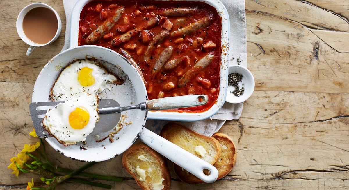Wurstauflauf mit Bohnen und Speck in Tomatensoße, serviert mit krossen Spiegeleiern und frisch getoasteten Brioche-Scheiben. 