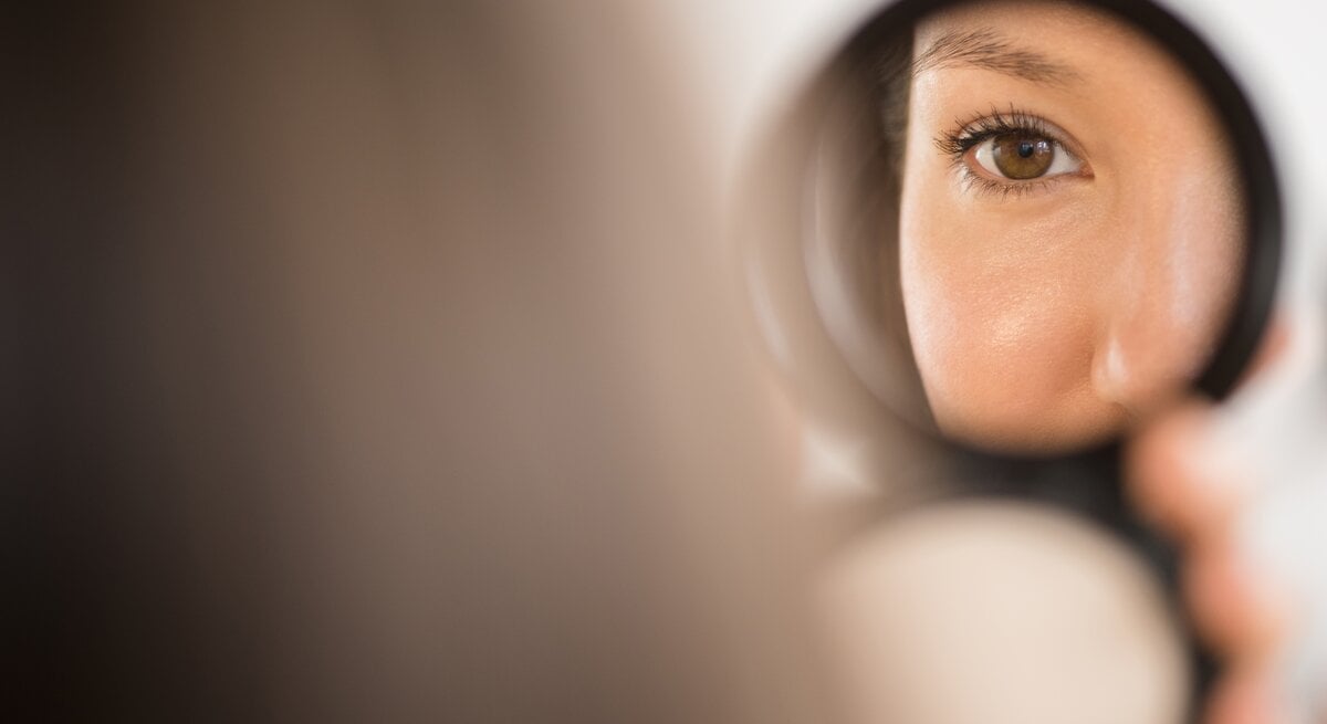 Eine Frau schaut in einen kleinen Handspiegel – nur ihr Auge und ihre Nase sind zu sehen.
