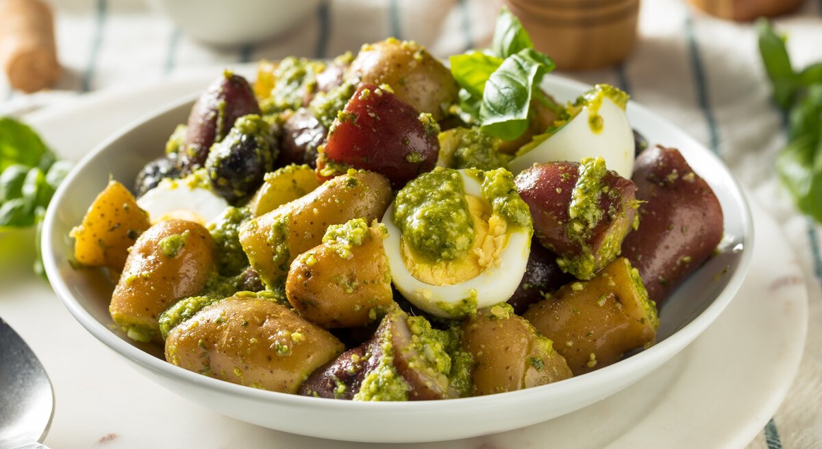 Kartoffelsalat mit grünem Pesto, Oliven und Ei in weißer Salatschüssel auf weißem Anrichteteller auf blau-weißem Tuch. Daneben Basilikumblätter. Darüber Pfeffermühle aus Holz. Frontalaufnahme.